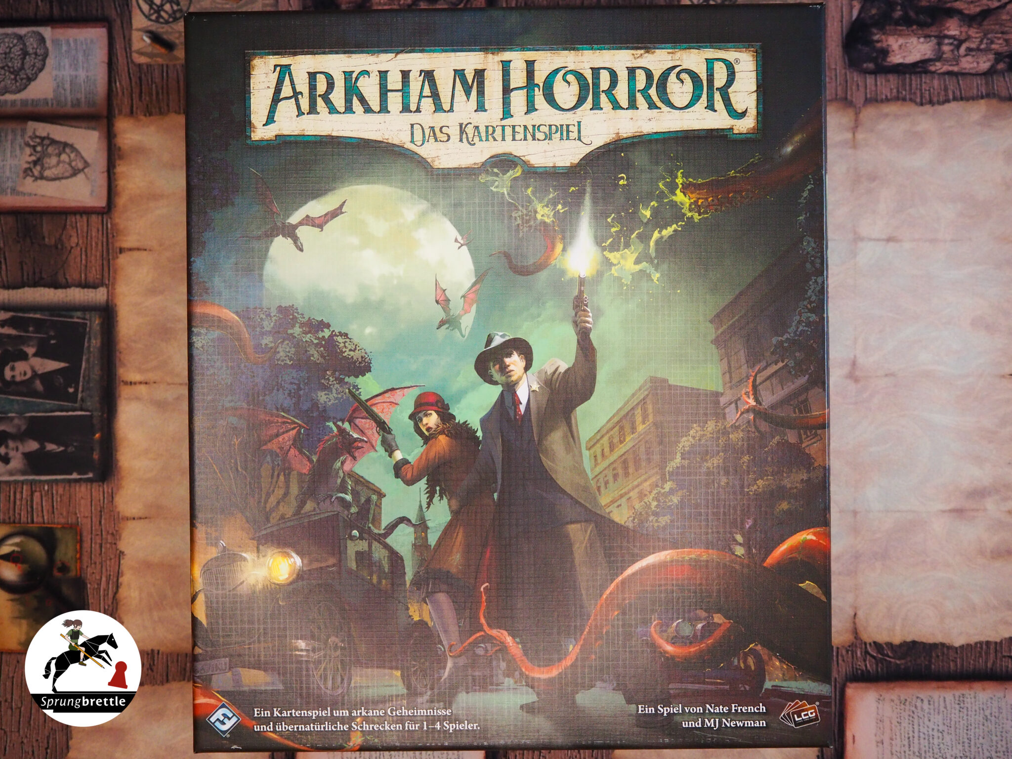 Die Spielschachtel von Arkham Horror Das Kartenspiel liegt mittig auf einem mittelbraunen Hintergrund. Sie ist vornehmlich in dunklem grün, schwarz und braun gehalten. Rechts erheben sich mehrstöckige, eckige Gebäude mit vielen Fenstern, Davor auf der Straße stehen eine weibliche und eine männliche Person in abwehrender Haltung mit alten Revolvern in der Hand. Die Person im Vordergrund feuert die Waffe ab und ein Lichtblitz erscheint. An verschiedenen Stellen des Bilds sind fliegende Ungeheuer mit roten, durchschimmernden Flügeln. Vom Rand aus erstrecken sich braun, orangene Tentakeln Richtung Personen in der Mitte. Deckbau, Wendy, Roland, Daisy