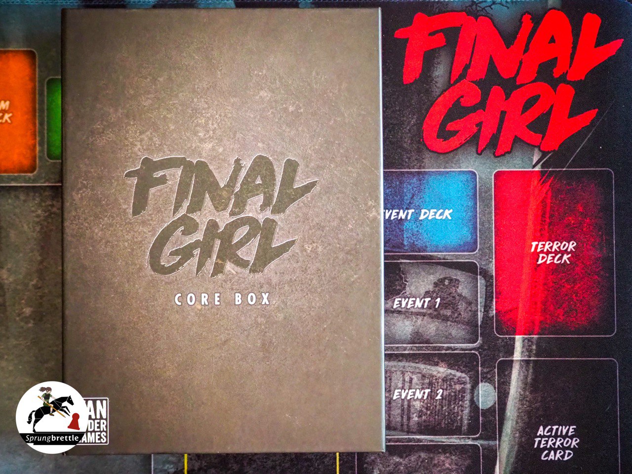 Der Hintergrund zeigt die dunkelgraue Spielmatte von Final Girl. Rechts oben im Bild steht der Titel Final Girl in dicken, roten Großbuchstaben. Außerdem sind weiß umrundete Kartenbereiche zu erkennen mit weißen Beschriftungen. Links der Mitte liegt die Grundbox in grau mit dunkelgrauer Titelschrift in der Mitte.