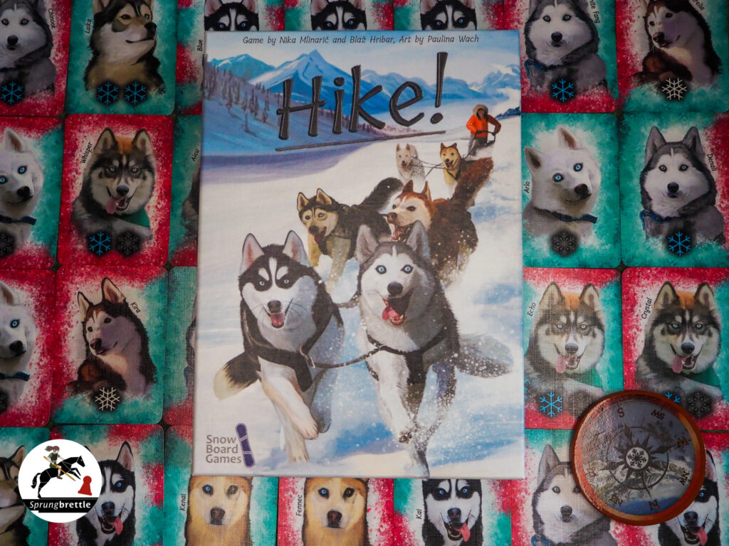 Das Kartenziehspiel Hike liegt mittig. Das Cover zeigt eine Schneelandschaft und sechs motivierte Huskies. Die Huskies haben weit offene Augen in blau und braun. Jeder Husky hat eine andere Farbe. Schwarz-weiß, grau-weiß, braun-weiß oder ganz weiß. Ihre Ohren sind spitz. Sie tragen ein Zuggeschirr und ziehen ein Person auf einem Schlitten hinter sich her. Die Person trägt eine Fellmütze, hat schulterlange, schwarze Haare und eine knallorangene Jacke. Im Hintergrund der Spielschachtel sind nebeneinander und untereinander über das komplette Bild Huskykarten aufgereiht.