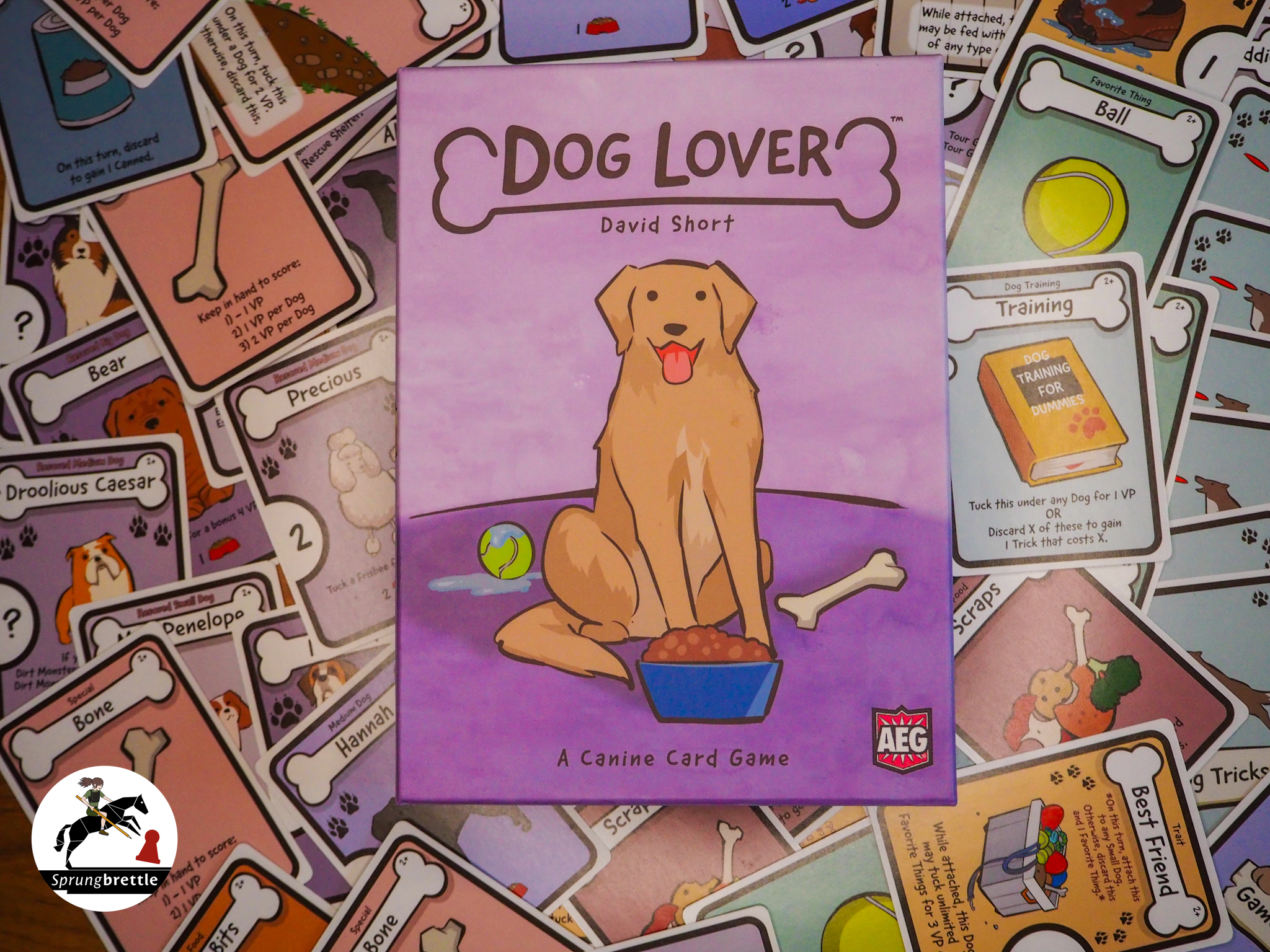 Die Spielschachtel des Sammelspiels Dog Lover liegt mittig auf einem zusammelgewürfelten Haufen der Spielkarten. Die Spieleschachtel ist im unteren Drittel dunkelrosa und darüber rosa. Mittig sitzt ein hellbrauner, hechelnder Hund mit Schlappohren. Neben ihm liegt ein gelber Tennisball, ein beiger Knochen und eine dunkelblaue Schüssel mit braunem Futter. Im oberen Drittel ist der Titel Dog Lover in dunkler Schrift in Großbuchstaben zu sehen.