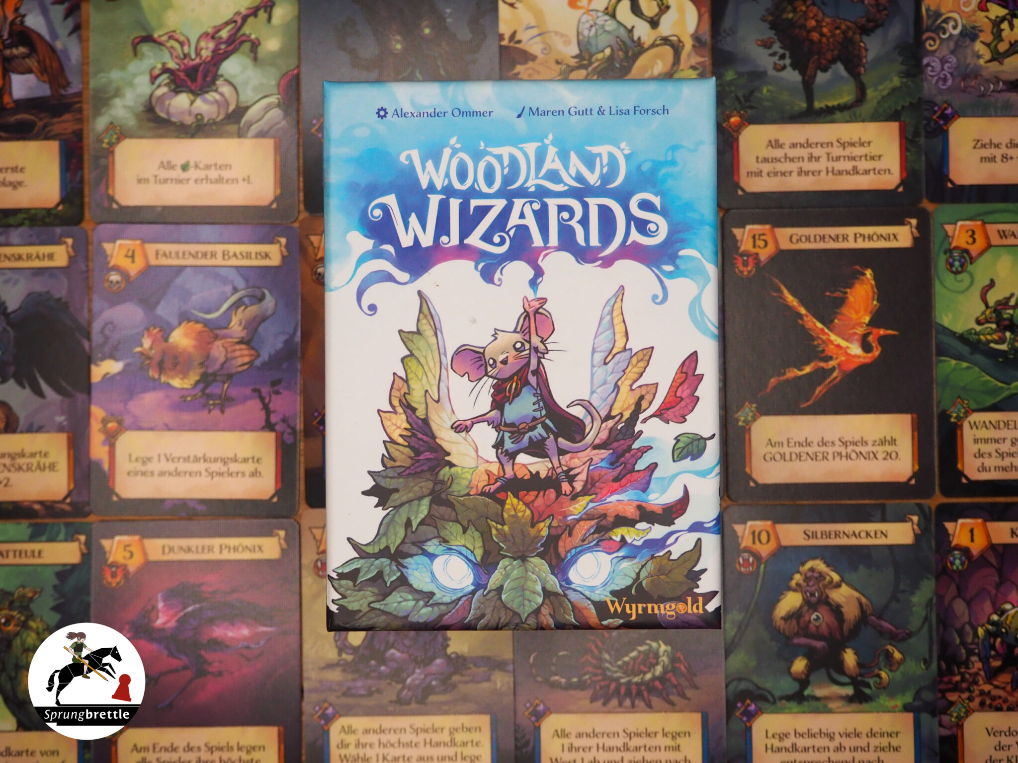 Kartenspiel Woodland Wizards ist mittig zu sehen. Im Hintergrund in Reihen und Spalten verschiedene Spielkarten aufgereiht.